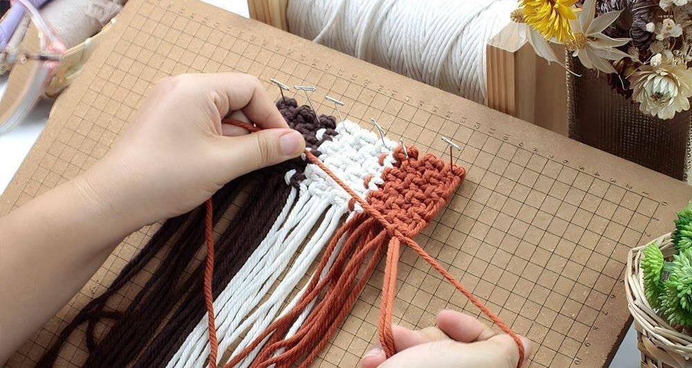 Hands tying macrame knots on a wooden cutting mat with natural fiber cords — the beginning of a handmade craft project.