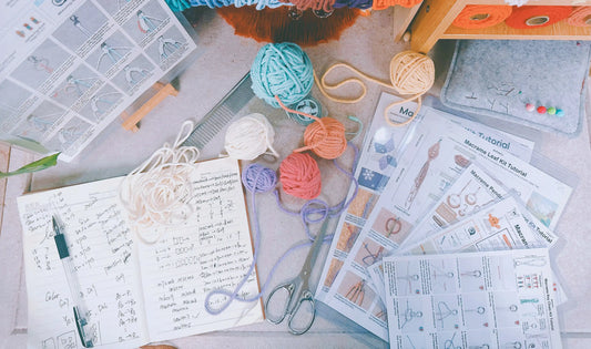 BUFANTL macrame kit development process — planning pages with cord measurements, colour codes and knot counts, surrounded by yarn balls, scissors and printed tutorials