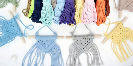 Top-down photo of a beginner macrame kit: four finished mini wall hangings with diamond openwork triangle patterns and soft fringe on small wooden dowels, beside coils of macrame cord in dozens of colors on a clean white background